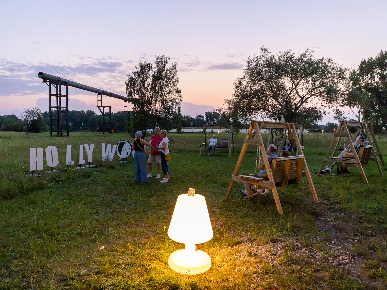 Impression der Veranstaltungsreihe HOLLYwow!Schaukelei des wowCLUBs. Auf dem Gelände stehen hölzerne Hollywoodschaukeln auf denen Menschen sitzen und mitteinander reden. Im Vordergrund steht eine überdimensionierte leuchtende Lampe.