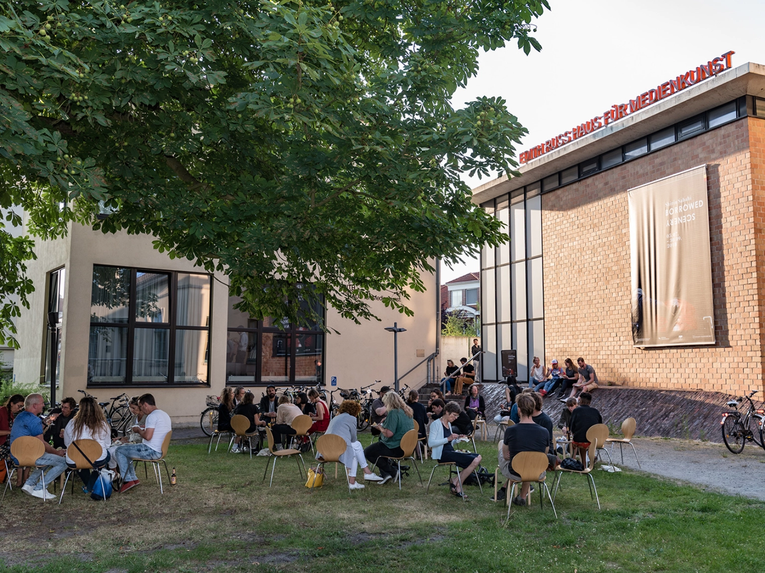Impression des Edith-Russ-Haus für Medienkunst in Oldenburg – Menschen sitzen auf der Grünfläche vor dem Gebäude.