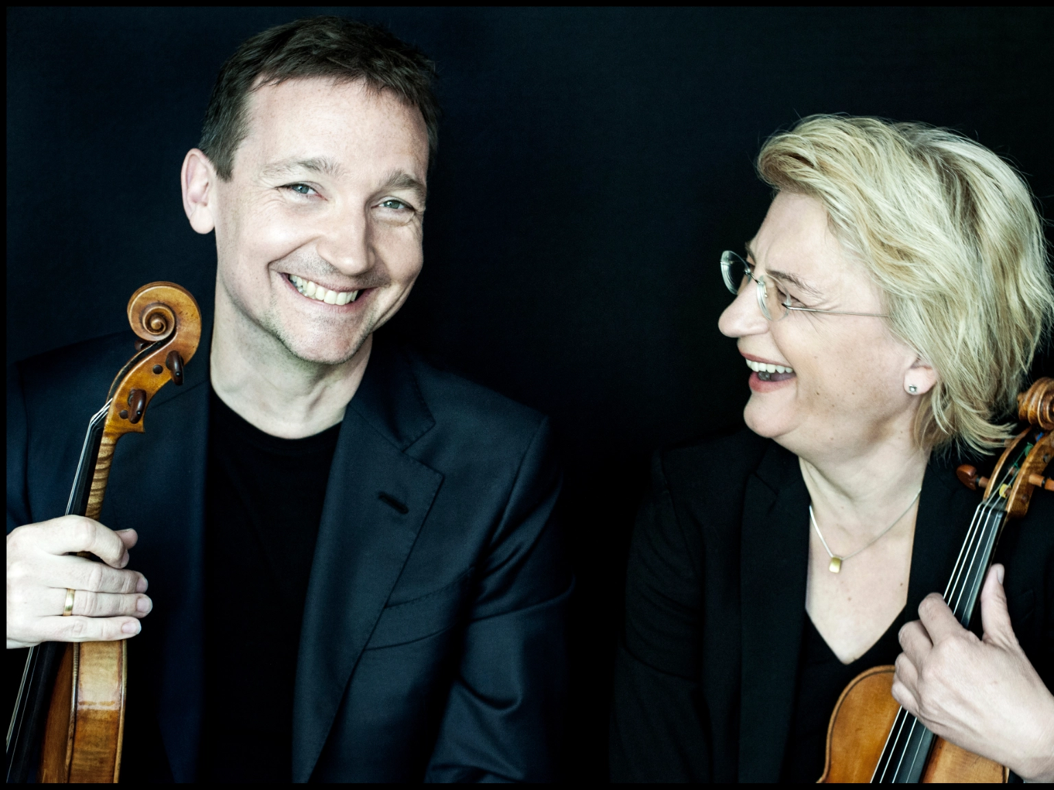 Die Künstlerische Leitung des Violinwettbewerbs: Antje Weithaas (rechts) und Oliver Wille (links). Beide halten eine Geige in der Hand. Oliver Wille lacht in die Kamera, Antje Weithaas schaut ihn lachend an.