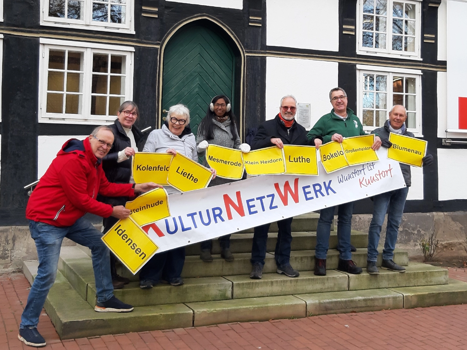 Gruppenbild: Die Engagierten des Vereins KulturNetzWerk Wunstorf freuen sich über die Förderung. Sie halten gemeinsam eine Banner mit dem Vereinsnamen hoch sowie kleinere gelbe Schilder mit den unterschiedlichen Namen de zu Wunstorf gehörenden Dörfer.