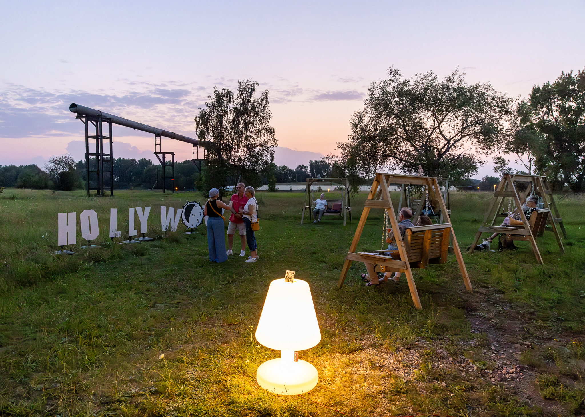 Impression der Veranstaltungsreihe HOLLYwow!Schaukelei des wowCLUBs. Auf dem Gelände stehen hölzerne Hollywoodschaukeln auf denen Menschen sitzen und mitteinander reden. Im Vordergrund steht eine überdimensionierte leuchtende Lampe.