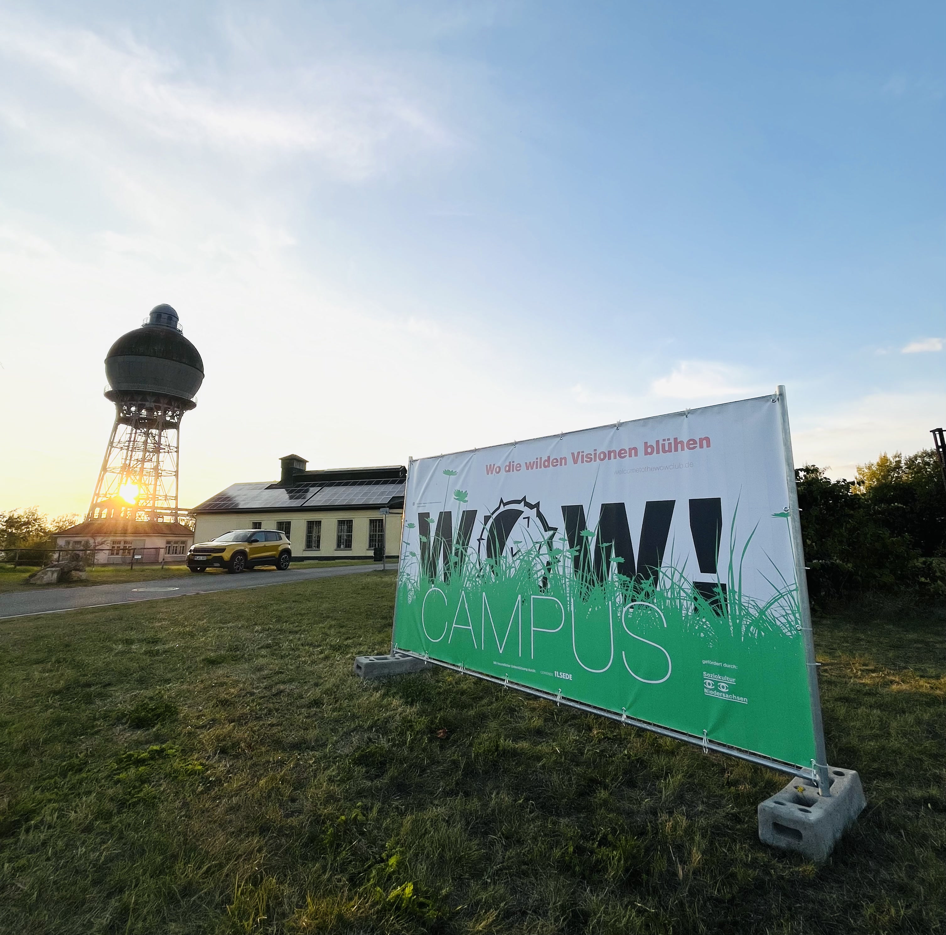 Impression des wow!CAMPUS mit dem Kugelwasserturm auf dem alten Hüttengelände in Ilsede, im Vordergrund steht ein Banner mit der Aufschrift wow!CAMPUS