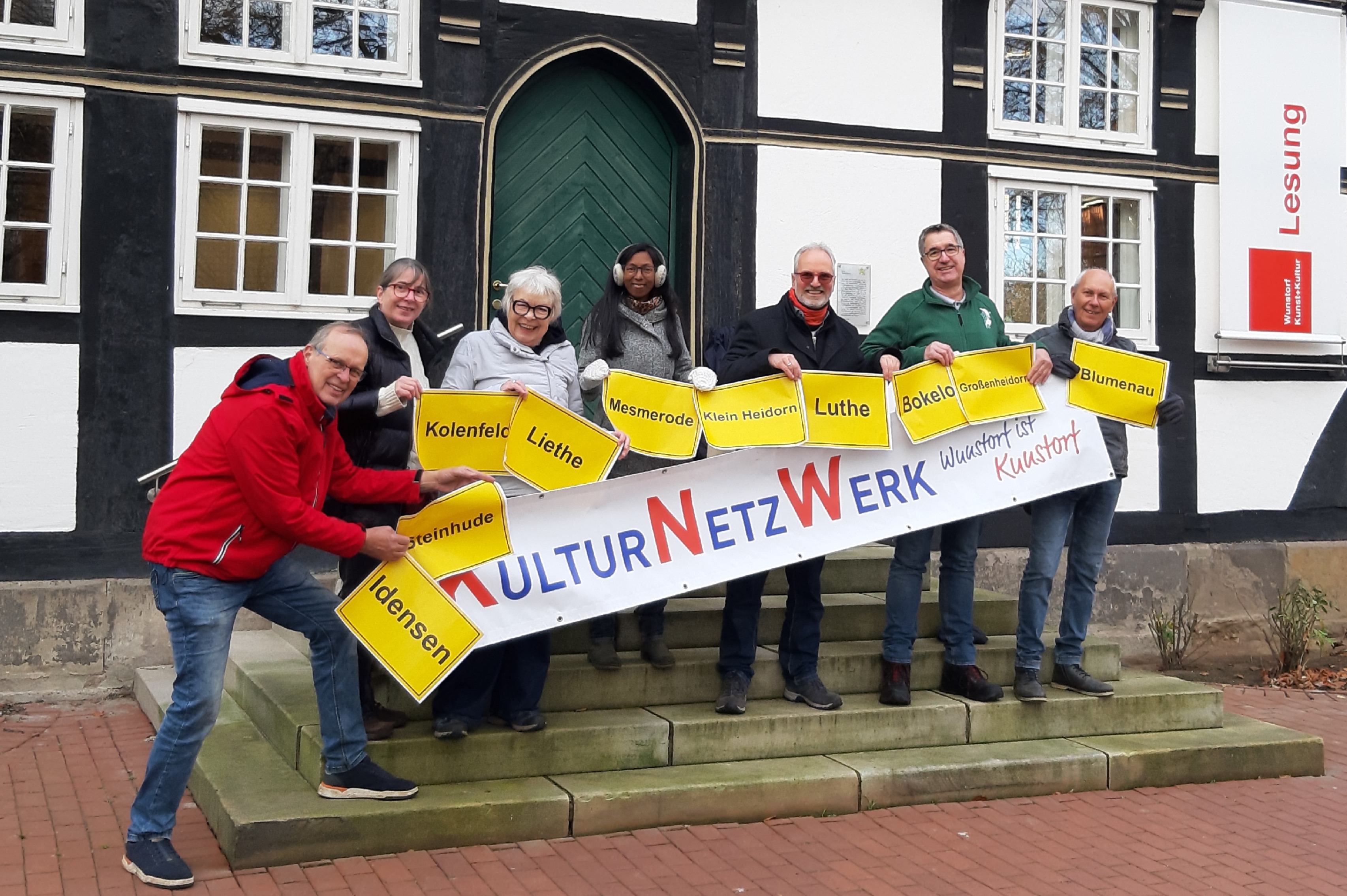 Gruppenbild: Die Engagierten des Vereins KulturNetzWerk Wunstorf freuen sich über die Förderung. Sie halten gemeinsam eine Banner mit dem Vereinsnamen hoch sowie kleinere gelbe Schilder mit den unterschiedlichen Namen de zu Wunstorf gehörenden Dörfer.